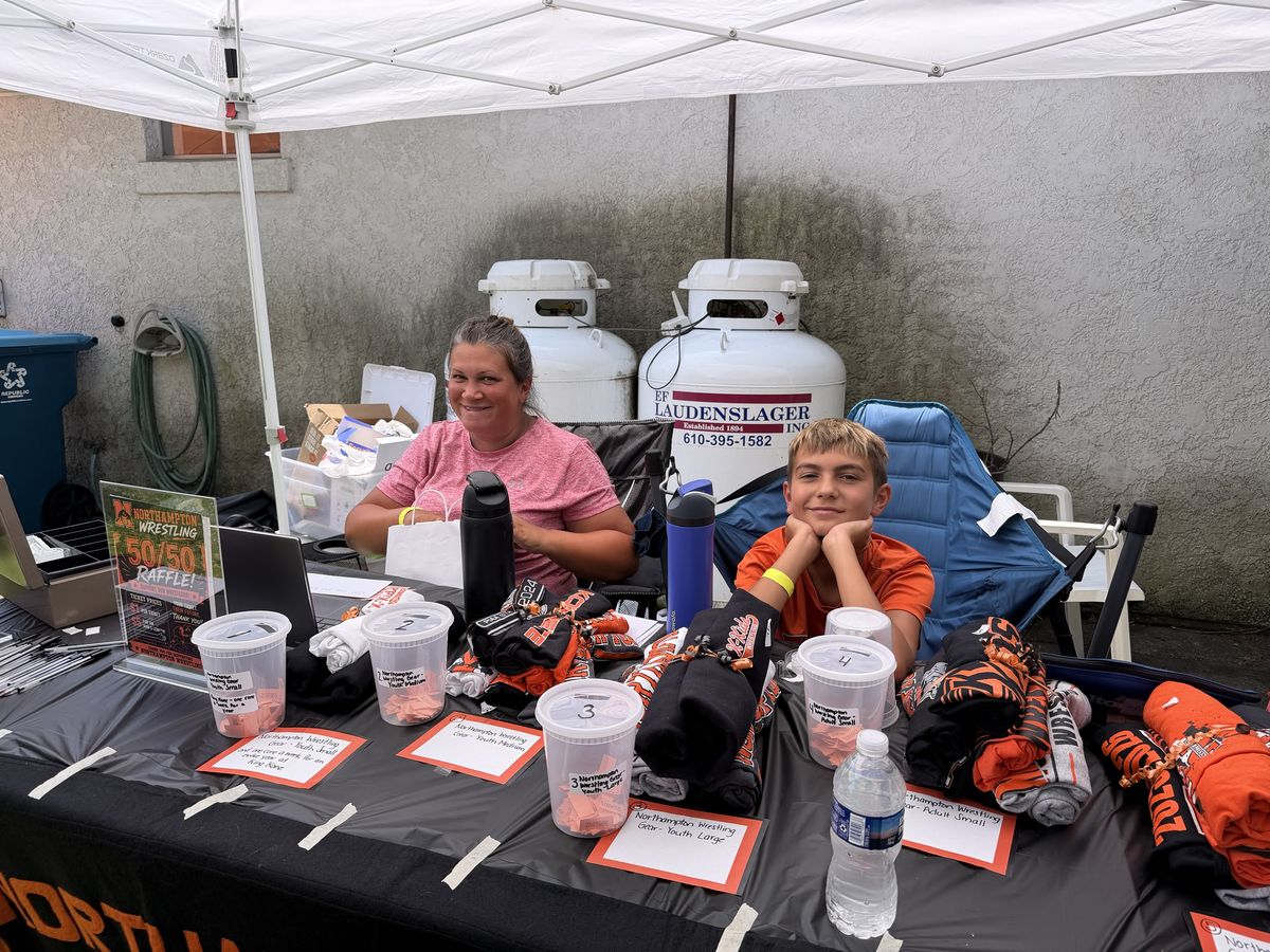 A volunteer and a young helper sit behind the 50/50 and Northampton Wrestling gear raffle table