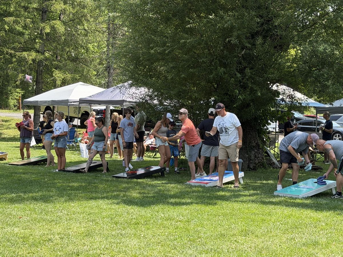 A row of players tosses bags at boards under the trees